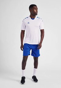 Vitt sportlinne med blå detaljer och logotyp, tillsammans med blåa shorts. Modellen bär svarta sneakers och vita strumpor. Rent, sportigt utseende.