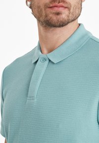 Light blue polo shirt with a textured pattern, featuring a classic collar and two-button placket. Fabric has a soft, woven appearance.