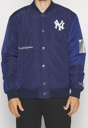 Mężczyzna w granatowej bomberce New York Yankees z zapięciem na guziki i czarnych spodniach, stojący na jednolitym tle.