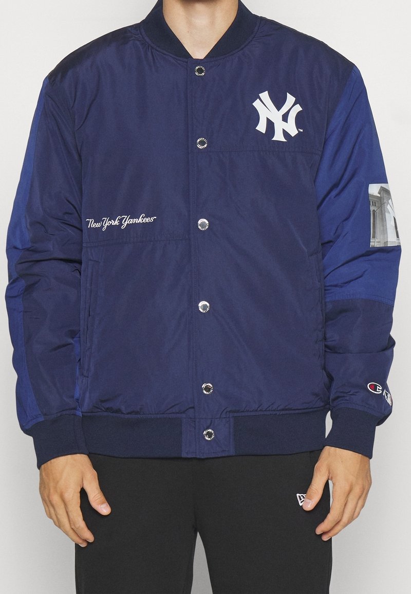 Homme portant une veste bomber bleu marine des New York Yankees avec fermeture à boutons et un pantalon noir, debout devant un fond uni.