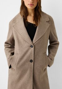 Donna che indossa un cappotto di lana beige con ampi rever e due bottoni neri, mani in tasca, su una maglia nera a coste.
