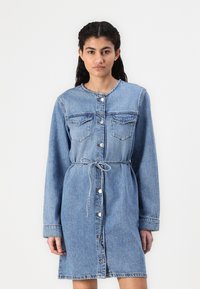 Svetlo modra denim obleka z okroglim ovratnikom, zapiranjem na gumb, dvema žepoma na prsih, vezanim pasom in dolgimi rokavi.