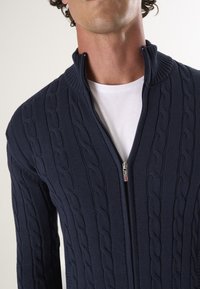 Navy Zip-Up-Pullover mit Kabelstrickstruktur, ausgestattet mit einem hohen Kragen und silbernem Reißverschluss. Über einem weißen Hemd getragen.