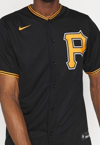 Uomo che indossa una maglietta nera da baseball con bordi arancioni, una grande "P" arancione sul petto e il logo swoosh arancione della Nike.