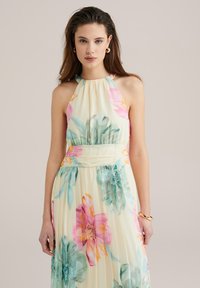 Flower maxi -mekko, jossa on halterneck-kaula-aukko, vaaleankeltainen kangas ja kirkkaan pinkkejä, oransseja ja teal-värisiä kukkia, sekä laskostettu hame.