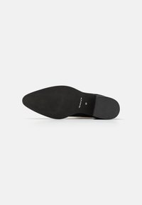 Suela de zapato negra con forma puntiaguda, superficie de goma texturizada y logo de la marca grabado. Incluye un ligero tacón y bordes limpios.