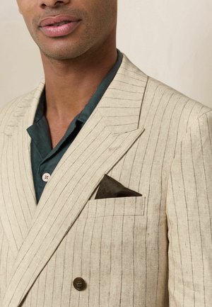 Man wearing a beige pinstripe blazer with dark brown pocket square over a dark green buttoned shirt, cropped from shoulders to lips.