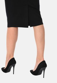 Scarpe nere con tacco alto, dalla finitura lucida, in morbido suede con punta affusolata e tacco a stiletto. Gonna di lunghezza Capri visibile.