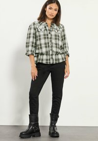 Mujer vestida con una blusa de cuadros verdes y blancos con botones y cuello con volantes, pantalones ajustados negros y botas negras hasta la mitad de la pantorrilla, de pie contra una pared blanca.