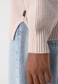 Pull rose clair en maille avec texture côtelée et poignets hauts. Porté sur un pantalon en denim bleu, avec une petite étiquette de marque sur la couture latérale.