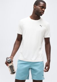 Άνδρας που φοράει λευκό t-shirt Puma και ανοιχτό μπλε σορτς Puma, κρατώντας ένα διάφανο μπουκάλι νερού με το λογότυπο "PUMA".