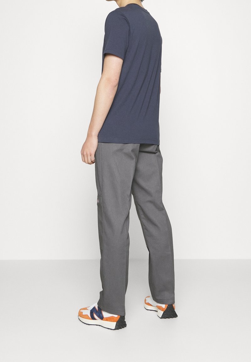 Personne debout de dos portant un t-shirt bleu marine, un pantalon gris et des baskets blanches avec des accents orange et bleu marine sur un fond blanc.