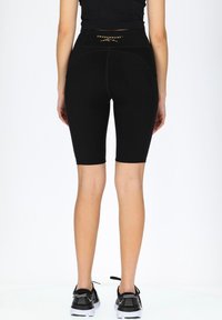 Svarta kompressionsshorts med hög midja, slät textur och minimala sömmar. Har en guldfärgad logotyp på ryggen, i kombination med svarta sneakers.