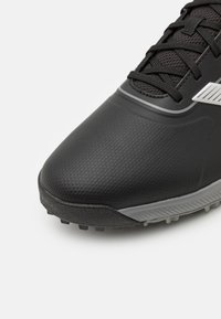 Zapatilla deportiva negra con parte superior texturizada, que presenta acentos grises, puntera reforzada y tapones moldeados en la suela exterior para mayor tracción.