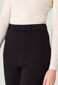Pantalons noirs ajustés avec une texture lisse, dotés d'une taille droite et de deux poches avant. Portés avec un haut à manches longues de couleur claire.