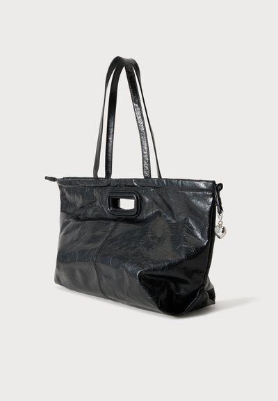 Sac cabas noir brillant avec double sangle d'épaule, poignée découpée rectangulaire et petit pendentif cœur argenté sur la fermeture éclair.