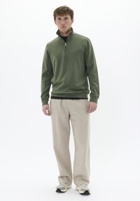 Pullover verde con zip al collo, texture liscia, abbinato a pantaloni beige a gamba larga e sneakers chiare. Modello in piedi su uno sfondo bianco.