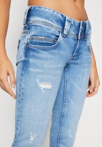 Los jeans de mezclilla azul claro presentan un ajuste slim, detalles ligeramente desgastados y una cinturilla de altura media con bolsillos delanteros y traseros.
