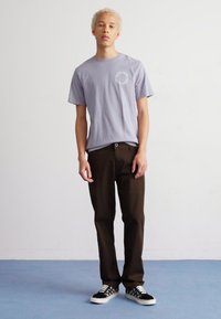 Camiseta de manga corta de color lila con un gráfico circular, combinada con pantalones marrón oscuro y zapatillas tipo slip-on a cuadros en blanco y negro.