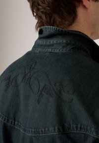 Chaqueta de mezclilla oscura con un patrón bordado en negro en la espalda, con un cuello clásico y una textura sutil.