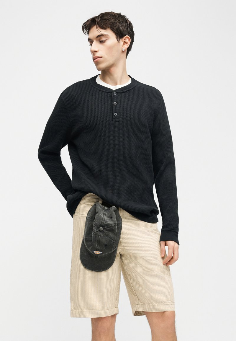Jonge man draagt een zwart lang mouw shirt met wafelstructuur en beige corduroy korte broek, hij houdt een zwarte pet vast in zijn voorzak en kijkt naar links.