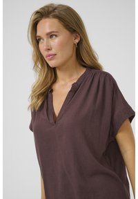 Blouse en lin marron à manches courtes avec un col en V et des détails plissés aux épaules. Texture douce et coupe décontractée.