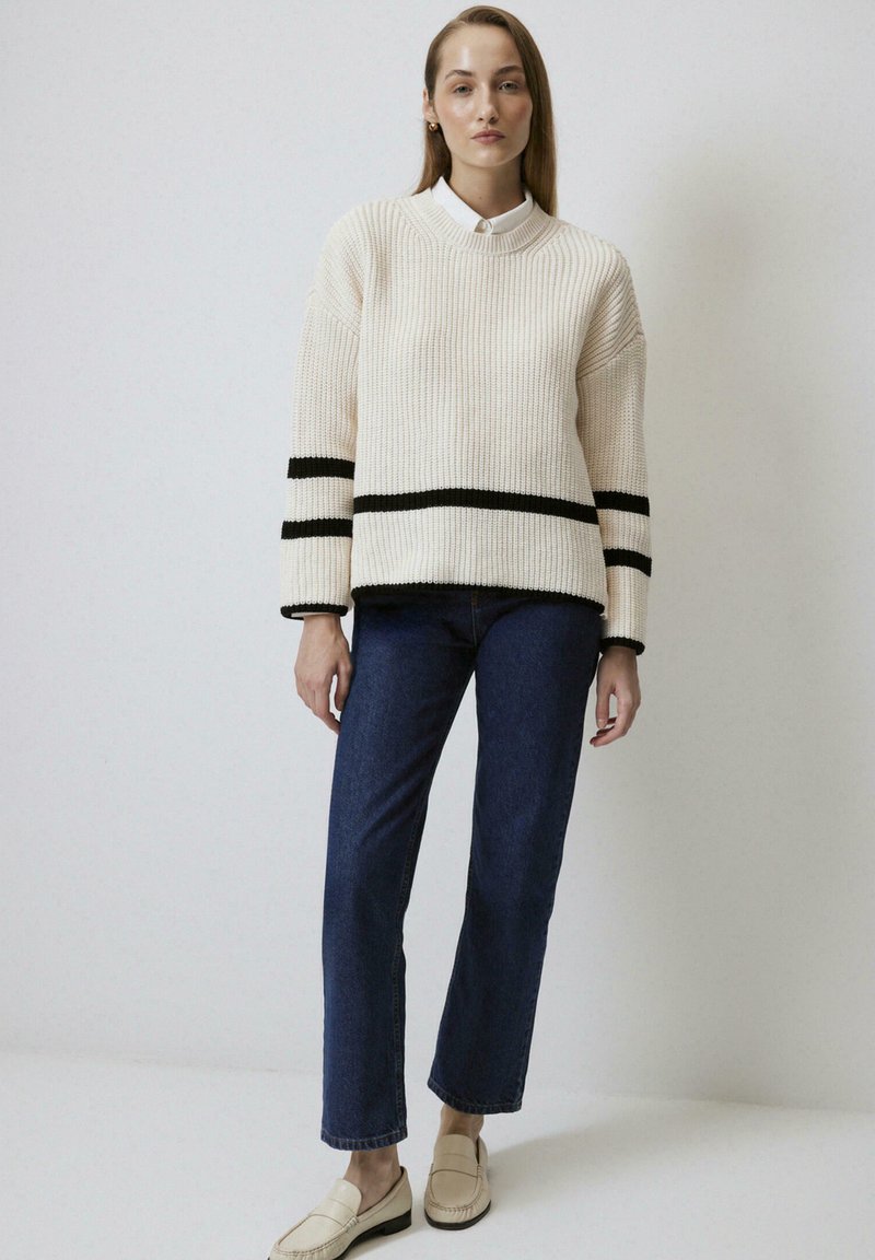 Gilet crème à côtes avec des rayures noires, col rond, épaules abaissées ; associé à un jean droit bleu et des mocassins de couleur claire.