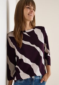Maglia a maniche lunghe viola e crema con un audace motivo ondulato, realizzata in morbido tessuto, abbinata a jeans di denim, posizionata davanti a una parete neutra.