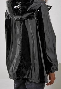 Chaqueta negra, brillante, impermeable, con capucha, puños elásticos y un ajuste holgado. Presenta una superficie brillante y un diseño de espalda recta.