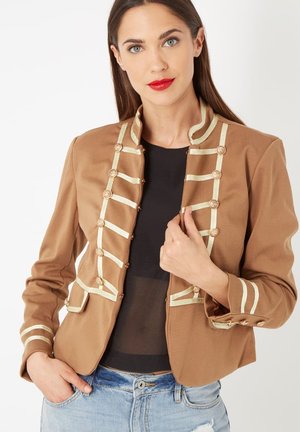 Mujer vistiendo una chaqueta militar de color arena con ribetes y botones dorados, blusa negra transparente y jeans claros desgastados, posando con una mano en la chaqueta.