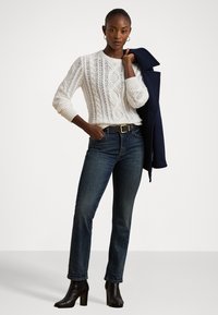 Sweater branco de malha com manga longa, jeans azul escuro e botas de salto pretas até o tornozelo; modelo segura um casaco azul-marinho sobre o braço.