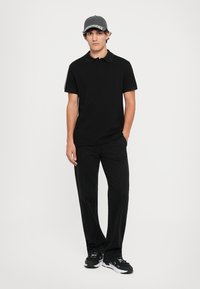 Black polo shirt with short sleeves, paired with black trousers featuring visible stitching. The model wears a grey cap and black sneakers.