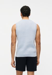 Άνδρας με σγουρά μαλλιά, που φοράει ανοιχτό γαλάζιο αμάνικο πουλόβερ με πλέξη καλώδιο και μαύρο σορτς, με την πλάτη γυρισμένη προς λευκό φόντο.