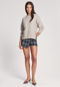 Beżowy sweter z dzianiny o prążkowanym wykończeniu, zestawiony z niebieskimi szortami w kratę. Modelka stoi w brązowych szpilkach, prezentując swobodny i casualowy strój.