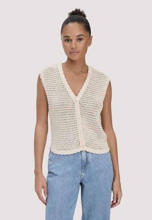 Frau trägt eine ärmellose, cremefarbene Häkelweste mit Knöpfen, kombiniert mit hellblauen, hoch taillierten Jeans, stehend vor einem schlichten Hintergrund.