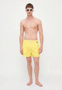 Jaunas vyras, dėvintis baltus saulės akinius ir geltonus maudymosi šortus, stovi basomis prieš baltą foną.