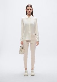 Μπουφάν από ελαφρύ κρεμ ύφασμα με κουμπιά, παντελόνι σε απαλό ροζ, μοντέρνα εφαρμοστή γραμμή, λευκά αθλητικά παπούτσια και μικρό ανοιχτόχρωμο τσαντάκι χειρός.