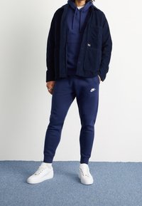 Navyblå cordfluwjacka över en navy hoodie och matchande sweatpants. Vita sneakers med snören och subtil branding på byxorna.