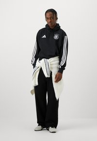 Czarna bluza z kapturem z białymi paskami Adidas i emblematem Niemiec, założony na talii oversize'owy biały sweter, czarne szerokie spodnie oraz szare buty.