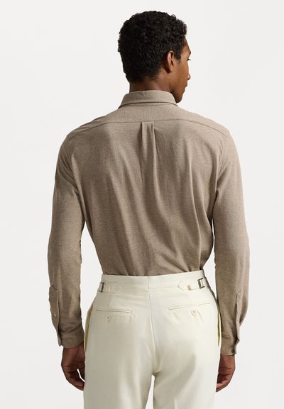 Chemise marron à manches longues avec col boutonné, tissu texturé et poignets à deux boutons, assortie à un pantalon crème avec des ajusteurs latéraux.