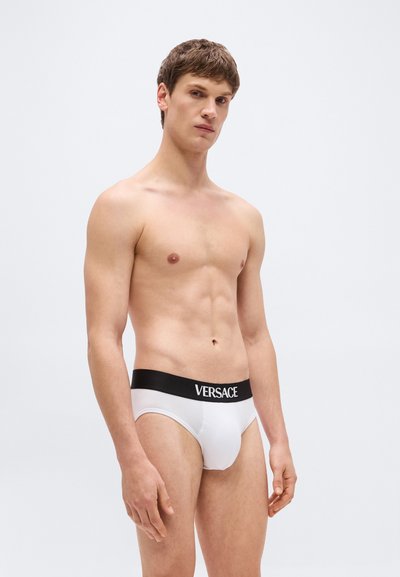 Jeune homme portant un slip Versace blanc avec une ceinture noire, debout contre un fond clair uni, légèrement tourné vers la gauche.