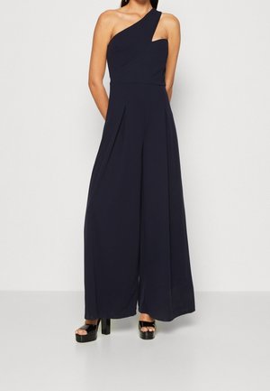 Woman wearing a navy blue one-shoulder wide-leg jumpsuit with pleats and black platform open-toe heels, standing against a plain background.