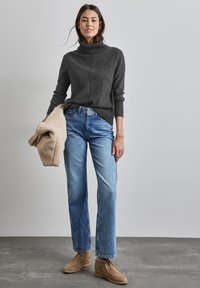 Grå rullkragsjumper, blå raka jeans och beige mockaskor. Håller ett texturerat beiget ytterplagg. Neutral bakgrund.