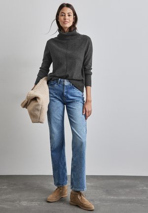 Pull en col roulé gris, jeans droits bleu, et chaussures en suède beige. Tient une pièce d'extérieur texturée beige. Fond neutre.