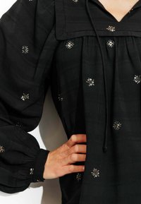 Blouse noire en tissu texturé, avec un décolleté carré, des manches ballons et des détails de broderie florale argentée partout.