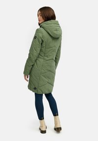 Olivengrønn, vattert lang kåpe med hette, med diamantmønster og en merkeetikett på venstre erme. Kombinert med mørke jeans.