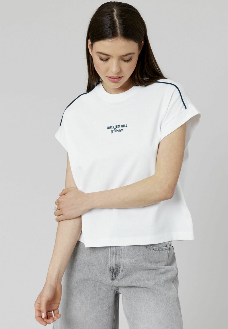 T-shirt blanc à manches courtes avec col rond, présentant le texte brodé en bleu marine "NOTTING HILL" sur la poitrine et des manches retroussées.
