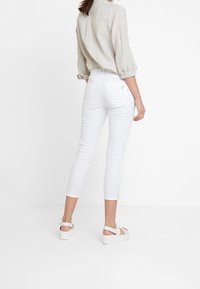 Mujer vestida con una blusa de manga larga beige, unos pantalones ajustados cortos blancos y sandalias de plataforma blancas, de pie contra un fondo blanco liso.