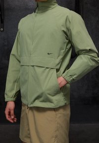 Chaqueta ligera de color verde claro, con cuello alto, cremallera frontal y puños elásticos, que presenta un logo negro en el pecho y un corte holgado.