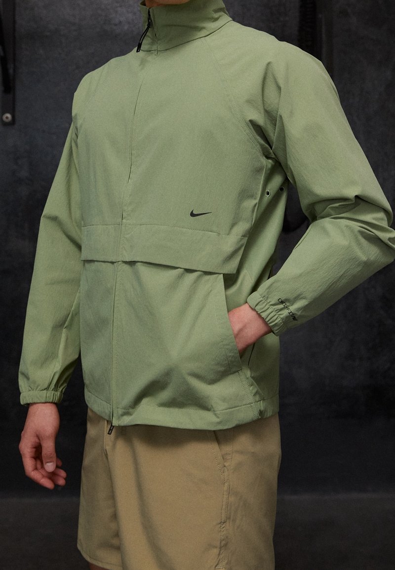 Chaqueta ligera de color verde claro, con cuello alto, cremallera frontal y puños elásticos, que presenta un logo negro en el pecho y un corte holgado.